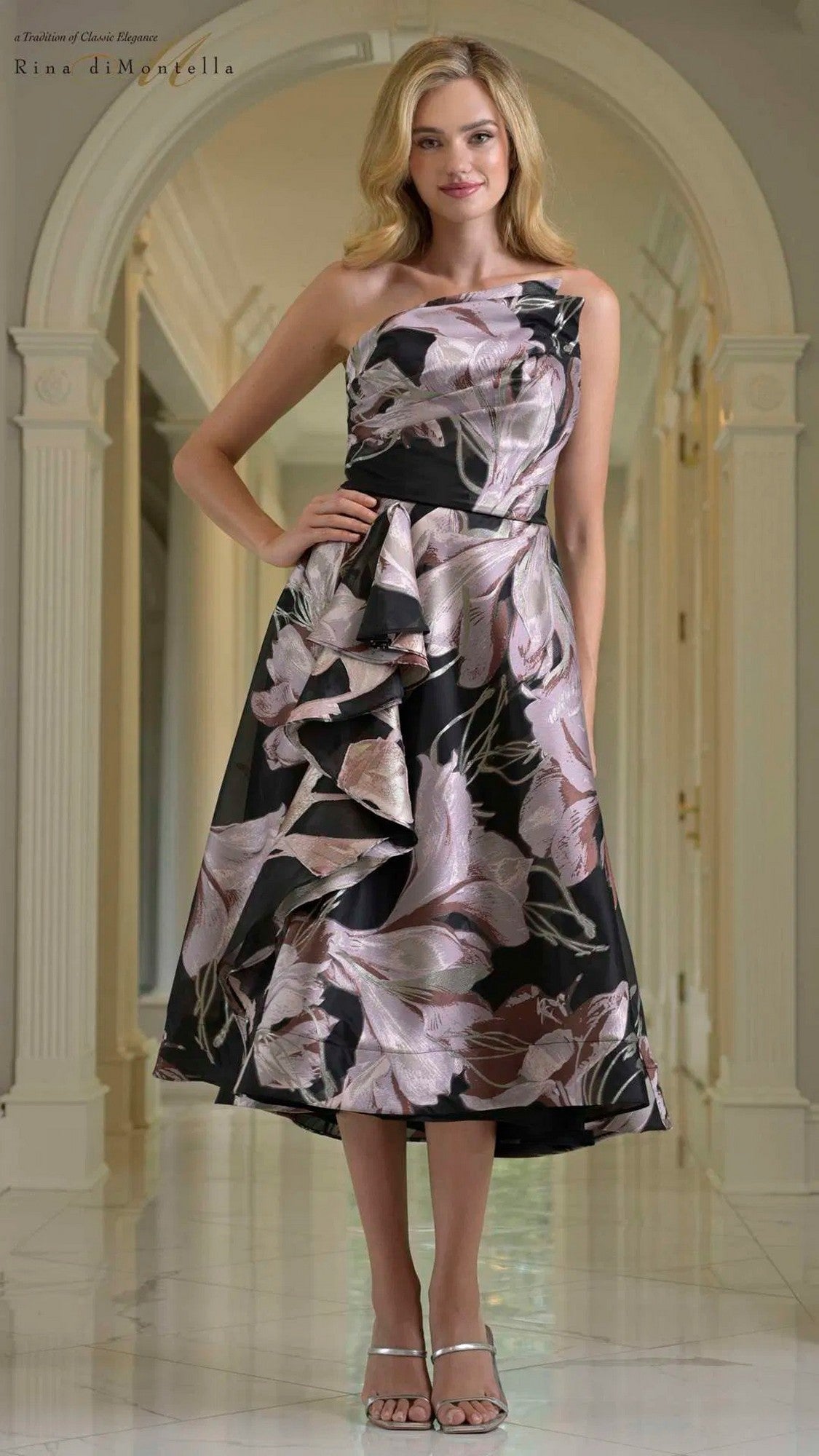 A woman stands in a hallway wearing the Rina Di Montella RD214 Knee Length Semi-Formal Dress in Black Mauve, featuring ruffle detail and a fit and flare silhouette with floral patterns, paired with strappy sandals.